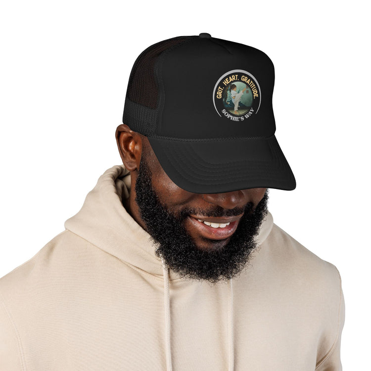 A black trucker hat with a circular graphic on the front panel. The graphic features a border with the words 'Grit. Heart. Gratitude. Sophie's Way' and a taekwondo tot doing a side kick in the center.