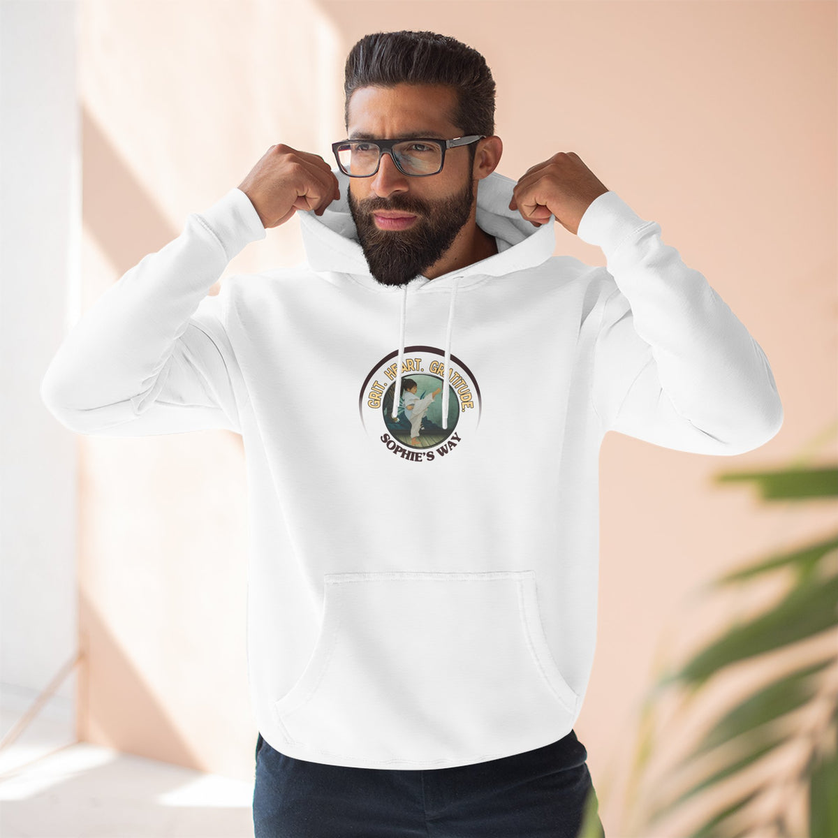 A white pullover hoodie with a circular graphic on the chest that reads 'Grit. Heart. Gratitude. Sophie's Way' around a globe design around a taekwondo tot doing a side kick. The hoodie features a drawstring hood, long sleeves, and a front kangaroo pocket.