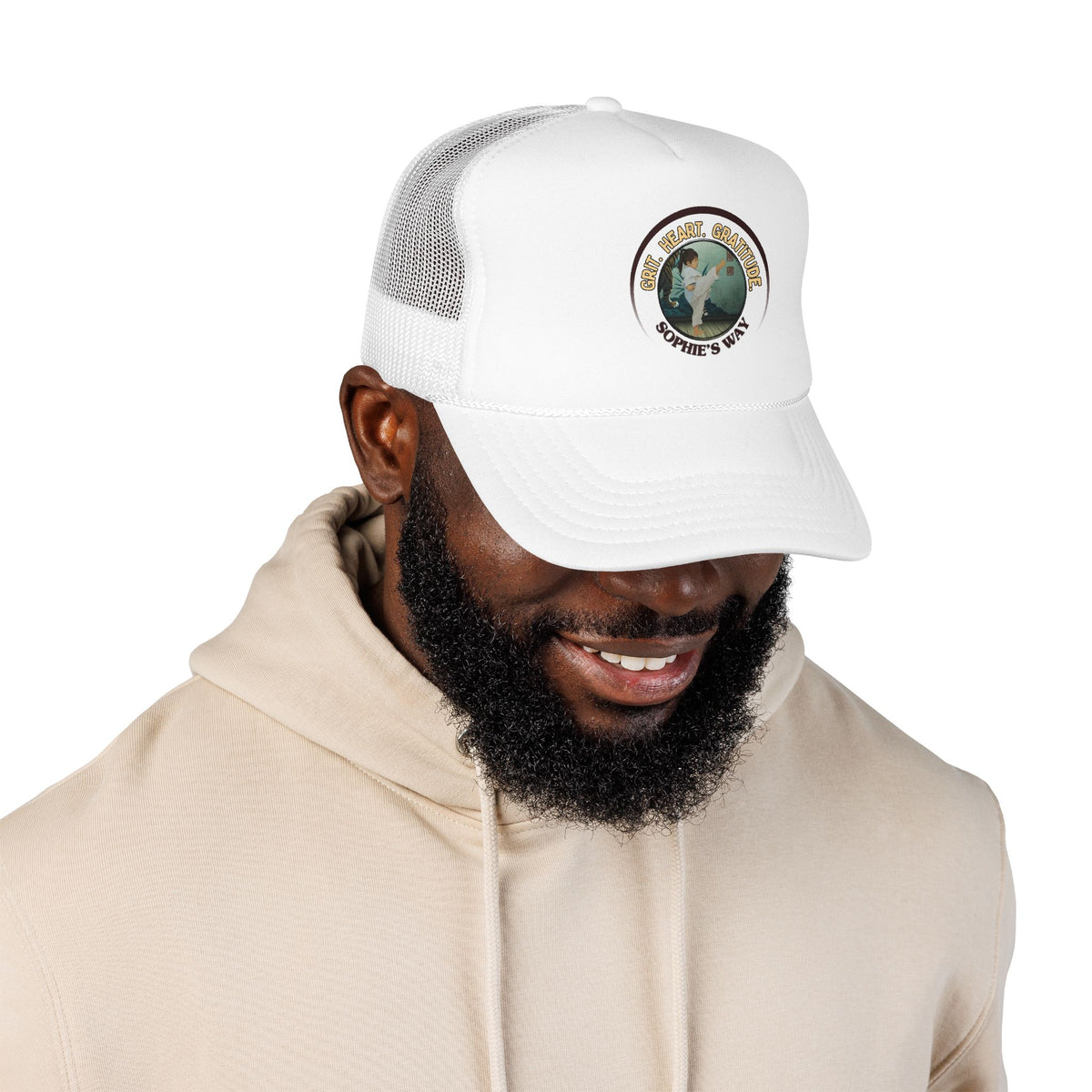 A white trucker hat with a circular graphic on the front panel. The graphic features a border with the words 'Grit. Heart. Gratitude. Sophie's Way' and a taekwondo tot doing a side kick in the center.