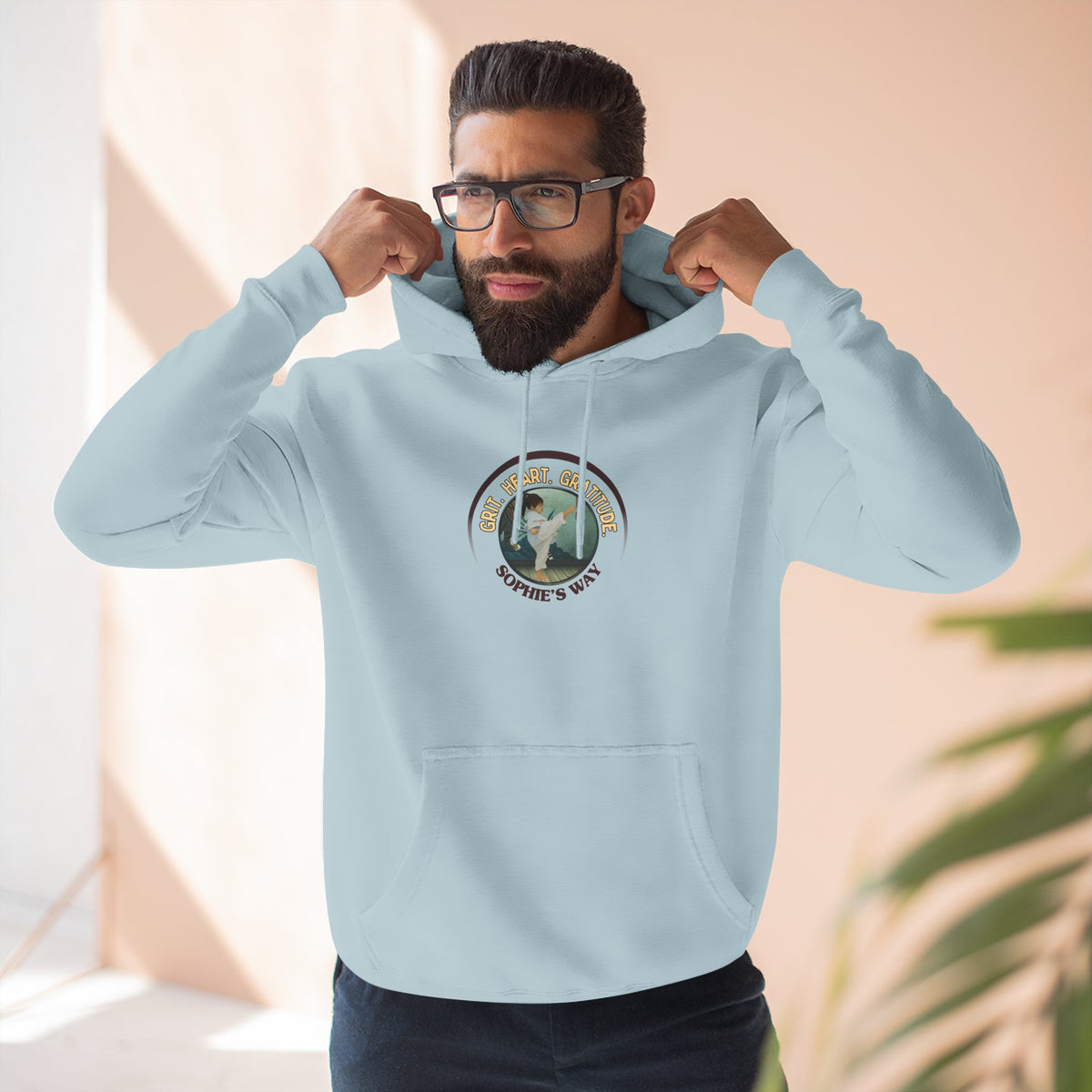 A blue mist pullover hoodie with a circular graphic on the chest that reads 'Grit. Heart. Gratitude. Sophie's Way' around a globe design around a taekwondo tot doing a side kick. The hoodie features a drawstring hood, long sleeves, and a front kangaroo pocket.