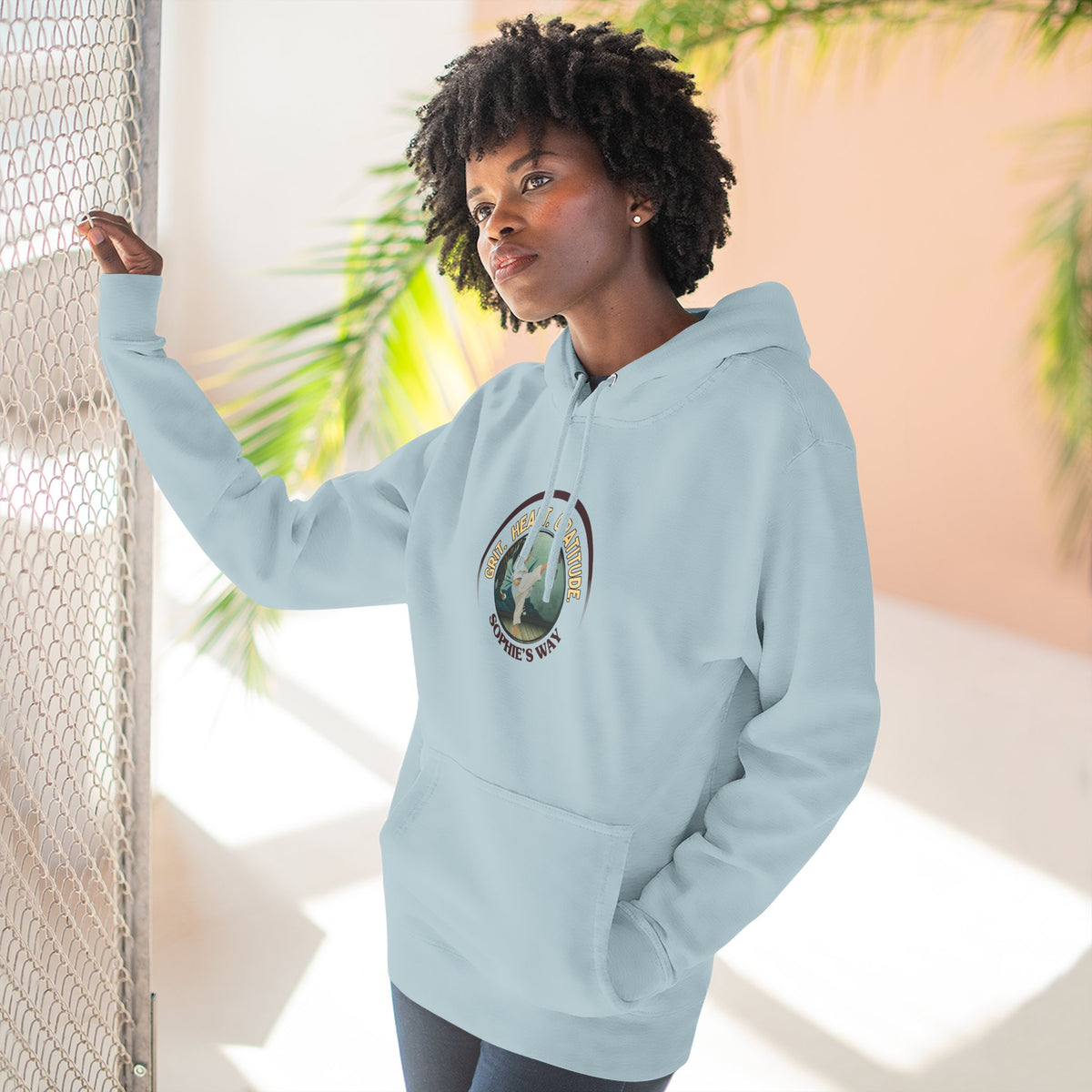 A blue mist pullover hoodie with a circular graphic on the chest that reads 'Grit. Heart. Gratitude. Sophie's Way' around a globe design around a taekwondo tot doing a side kick. The hoodie features a drawstring hood, long sleeves, and a front kangaroo pocket.