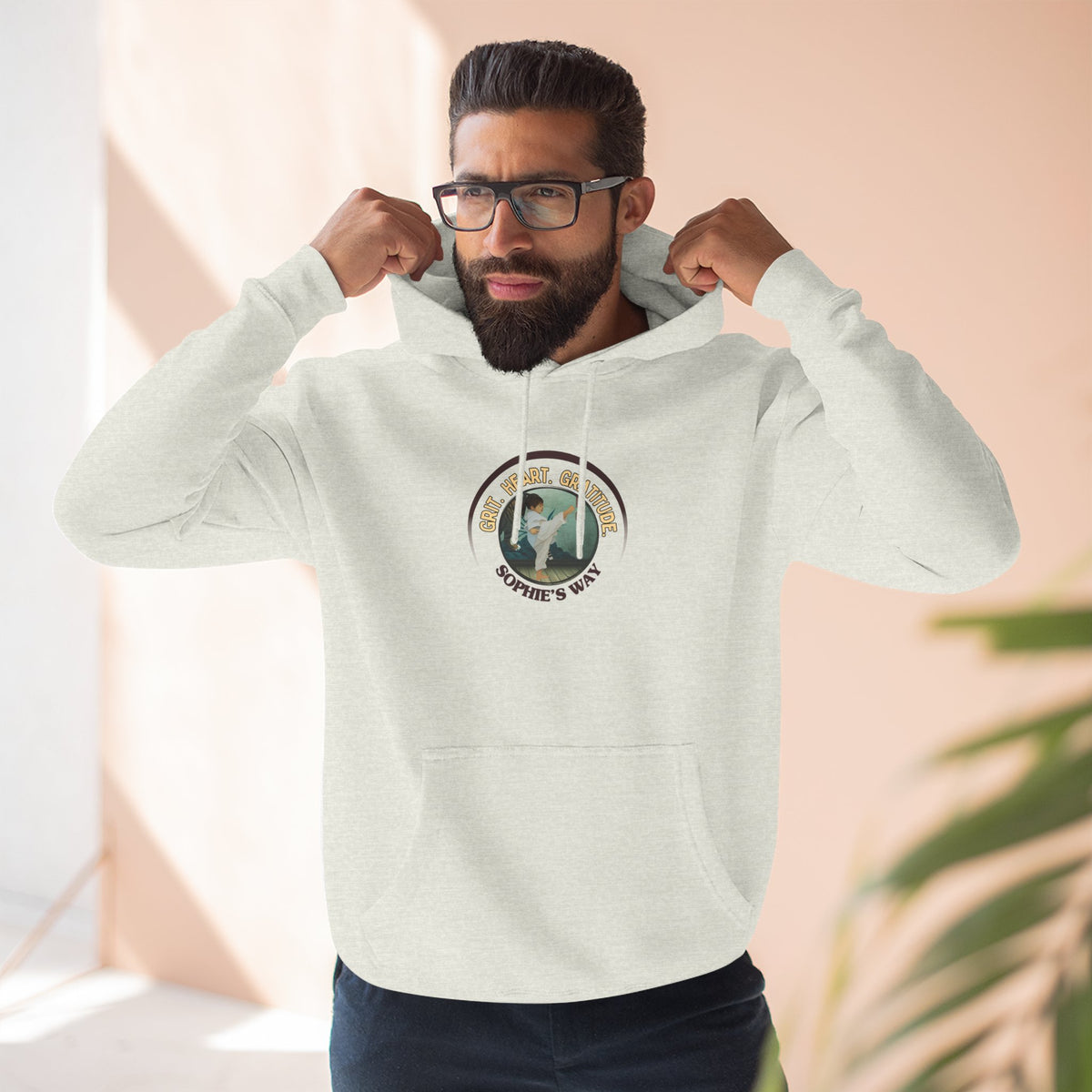 A oatmeal heather pullover hoodie with a circular graphic on the chest that reads 'Grit. Heart. Gratitude. Sophie's Way' around a globe design around a taekwondo tot doing a side kick. The hoodie features a drawstring hood, long sleeves, and a front kangaroo pocket.