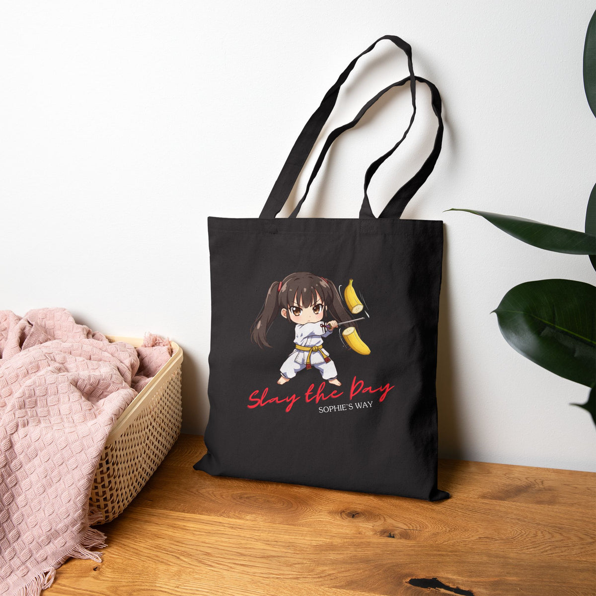 Slay the Day cotton canvas tote bag in natural, standing upright on wooden table with plant and basket — chibi taekwondo girl slicing a banana with a sword — "Slay the Day, Sophie's Way"