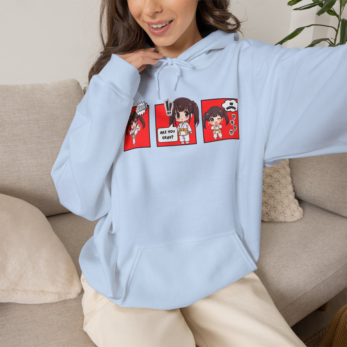 A light blue pullover hoodie with a red and multicolor anime-style graphic print on the chest of a taekwondo girl saying 'Aiyah!' then 'Are you okay?', and then 'Hi Mommy.'