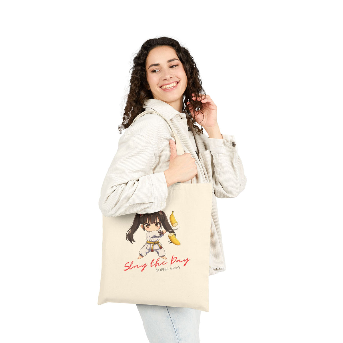 Slay the Day cotton canvas tote bag in natural, worn over shoulder by smiling woman — chibi taekwondo girl slicing a banana with a sword — "Slay the Day, Sophie's Way"