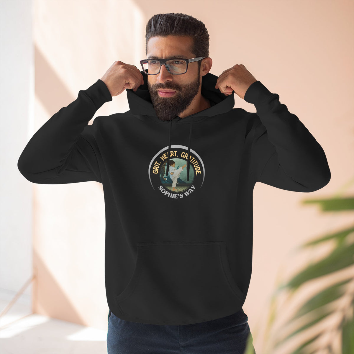 A black pullover hoodie with a circular graphic on the chest that reads 'Grit. Heart. Gratitude. Sophie's Way' around a globe design around a taekwondo tot doing a side kick. The hoodie features a drawstring hood, long sleeves, and a front kangaroo pocket.