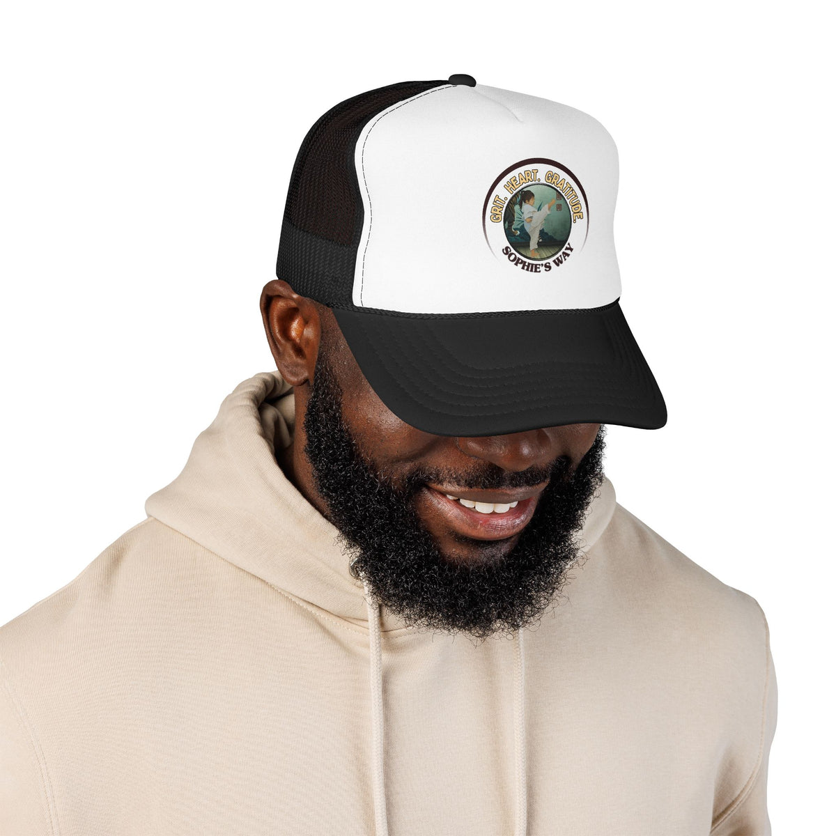 A black and white trucker hat with a circular graphic on the front panel. The graphic features a border with the words 'Grit. Heart. Gratitude. Sophie's Way' and a taekwondo tot doing a side kick in the center.