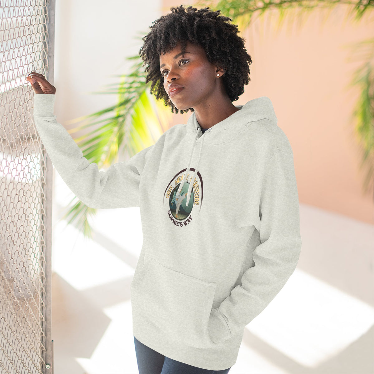 A oatmeal heather pullover hoodie with a circular graphic on the chest that reads 'Grit. Heart. Gratitude. Sophie's Way' around a globe design around a taekwondo tot doing a side kick. The hoodie features a drawstring hood, long sleeves, and a front kangaroo pocket.