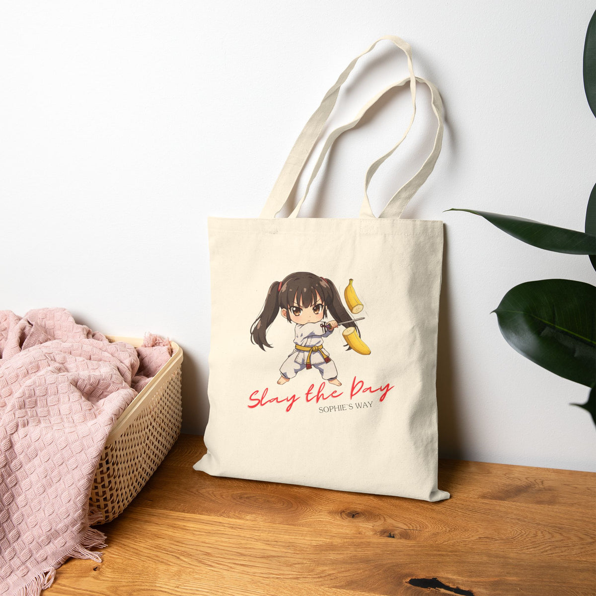 Slay the Day cotton canvas tote bag in natural, standing upright on wooden table with plant and basket — chibi taekwondo girl slicing a banana with a sword — "Slay the Day, Sophie's Way"