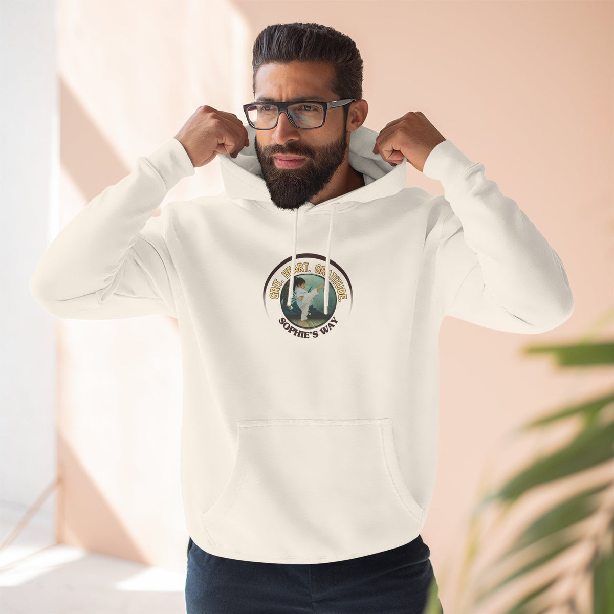 A sandshell pullover hoodie with a circular graphic on the chest that reads 'Grit. Heart. Gratitude. Sophie's Way' around a globe design around a taekwondo tot doing a side kick. The hoodie features a drawstring hood, long sleeves, and a front kangaroo pocket.