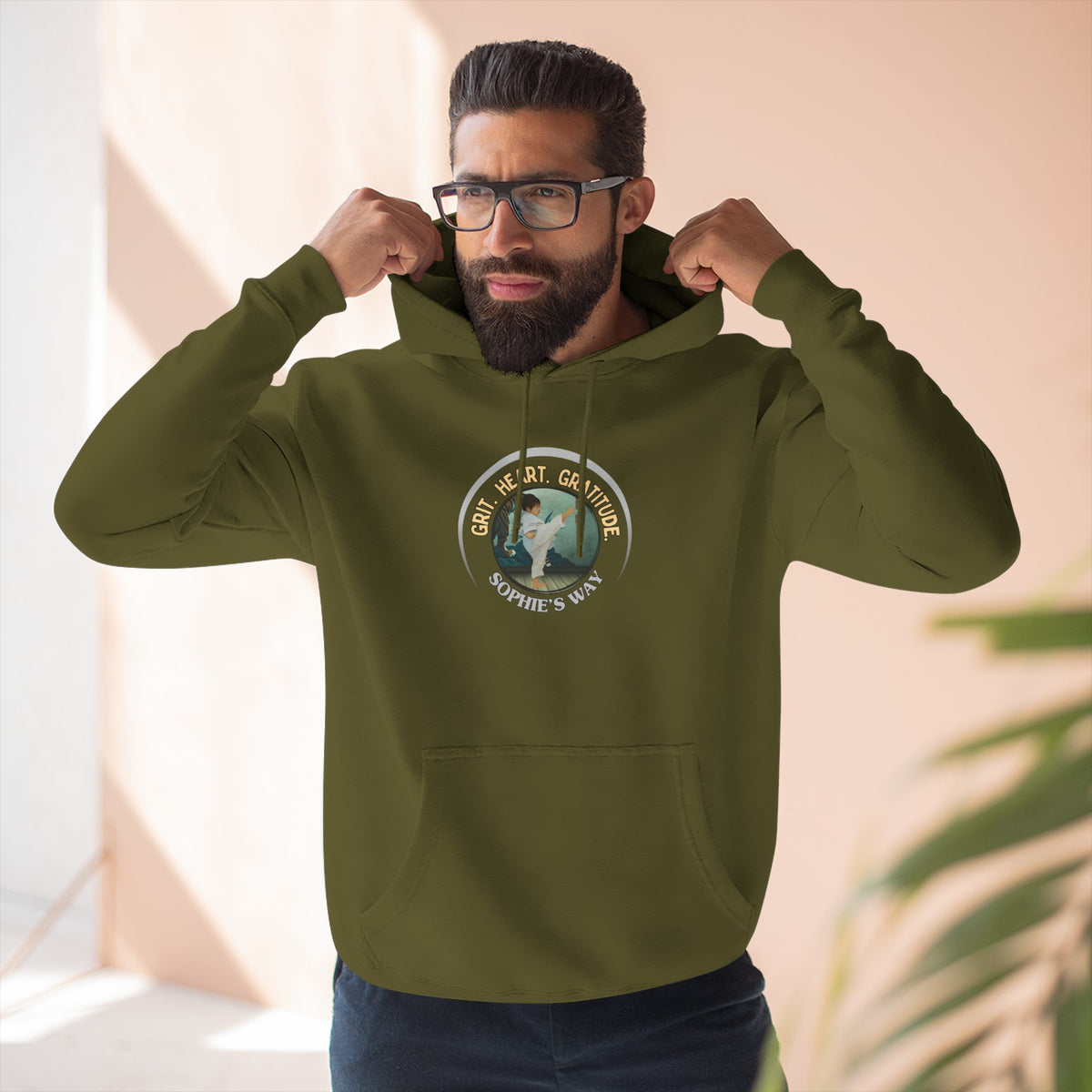 A army green pullover hoodie with a circular graphic on the chest that reads 'Grit. Heart. Gratitude. Sophie's Way' around a globe design around a taekwondo tot doing a side kick. The hoodie features a drawstring hood, long sleeves, and a front kangaroo pocket.