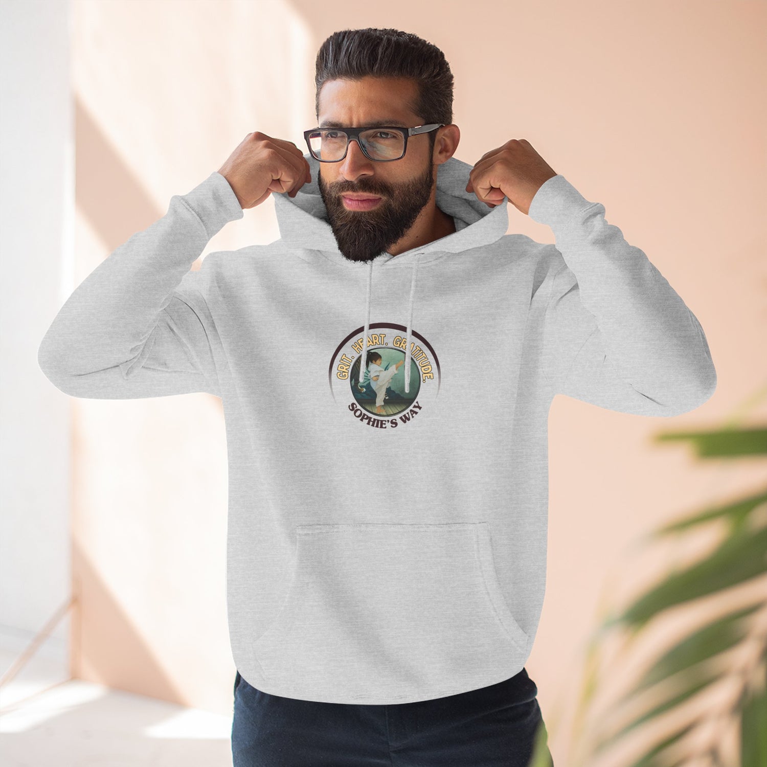 Grit, Heart, Gratitude fleece hoodie in heather grey, flat-lay — center-chest badge with taekwondo girl doing a side kick, martial arts studio background — "Grit, Heart, Gratitude, Sophie's Way"