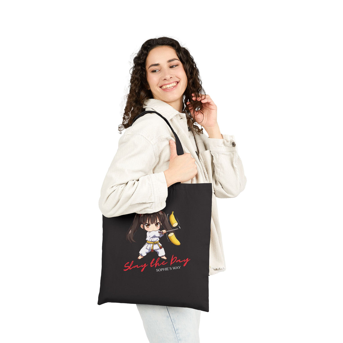 Slay the Day cotton canvas tote bag in black, worn over shoulder by smiling woman — chibi taekwondo girl slicing a banana with a sword — "Slay the Day, Sophie's Way"