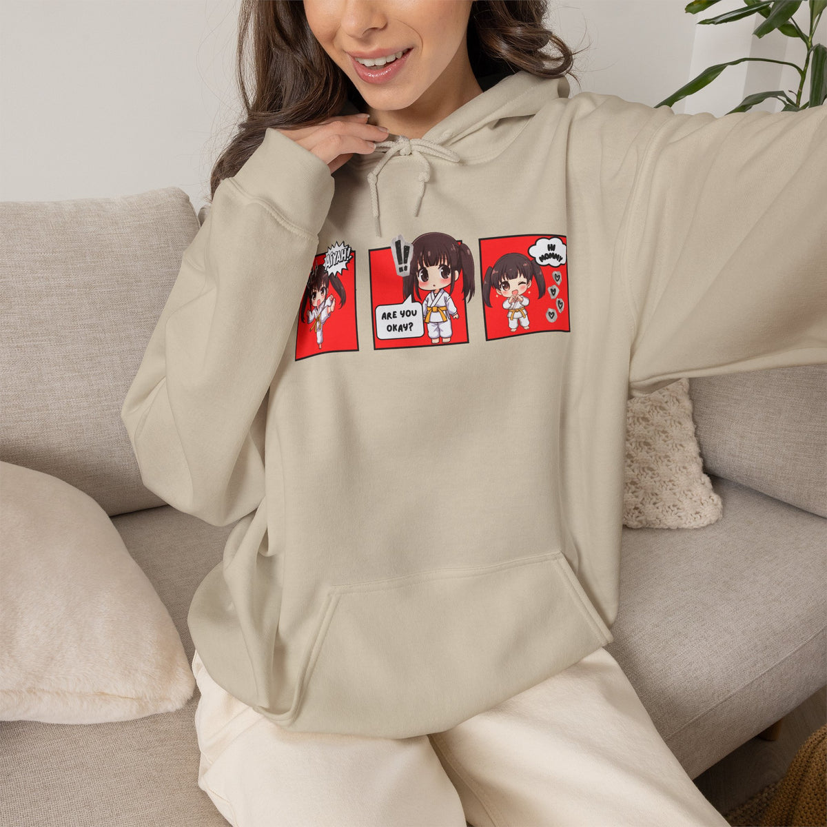 A sand color pullover hoodie with a red and multicolor anime-style graphic print on the chest of a taekwondo girl saying 'Aiyah!' then 'Are you okay?', and then 'Hi Mommy."