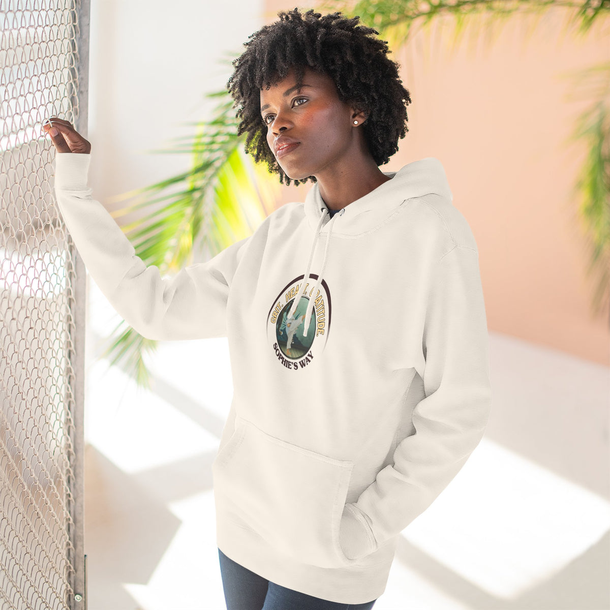 A sandshell pullover hoodie with a circular graphic on the chest that reads 'Grit. Heart. Gratitude. Sophie's Way' around a globe design around a taekwondo tot doing a side kick. The hoodie features a drawstring hood, long sleeves, and a front kangaroo pocket.